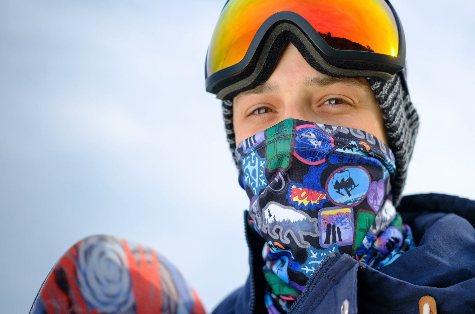 man snowboarding wearing goggles and vintage ski patch print neck gaiter